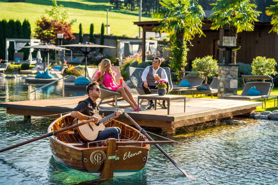 Paar entspannt auf Terrasse am Naturbadeteich mit Gitarrist im Boot